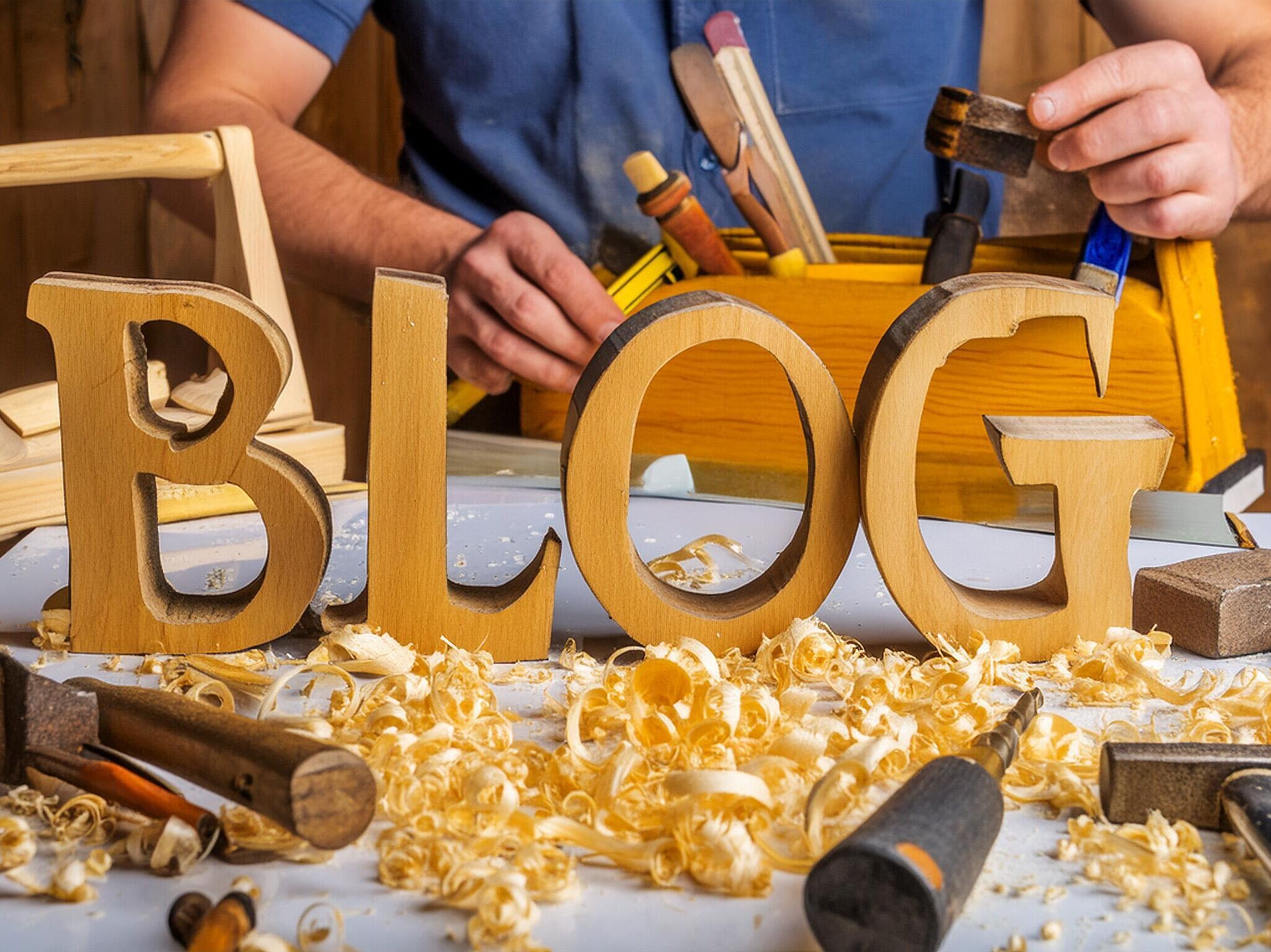 Auf einer Holzwerkbank stehen die Holzbuchstaben BLOG, ringsherum Werkzeuge zum Holz bearbeiten. Dahinter steht eine Person die aus einem Werkzeugkasten nach weiteren Werkzeugen zum Bearbeiten greift.